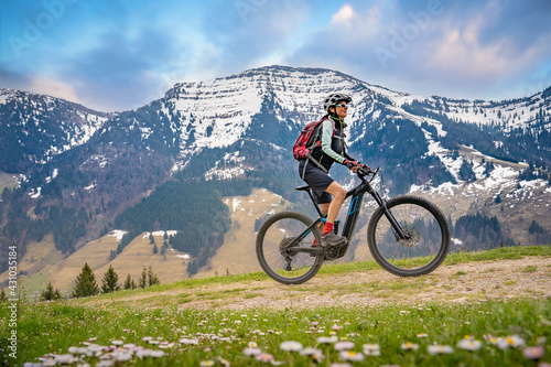 pretty mid age woman riding her electric mountain bike in early springtime in the Allgau mountains near Oberstaufen,below the spectacular snow capped mount HochgratNagelfluh chain