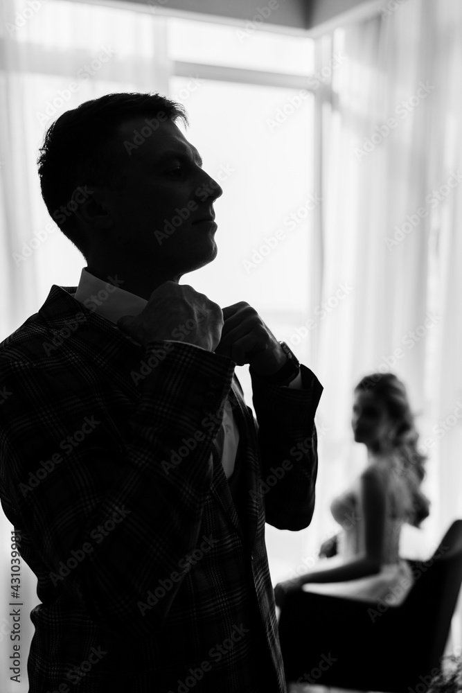 Male groom in a suit straightens a bow tie. The bride stands behind in a blurred background. Preparing a young couple for the wedding.