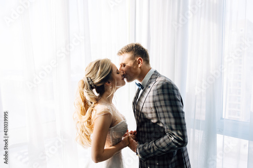 Handsome caucasian groom kisses european bride in white dress tender standing near window in sunny day. young wife and husband hold hands and have sincere feelings of love.