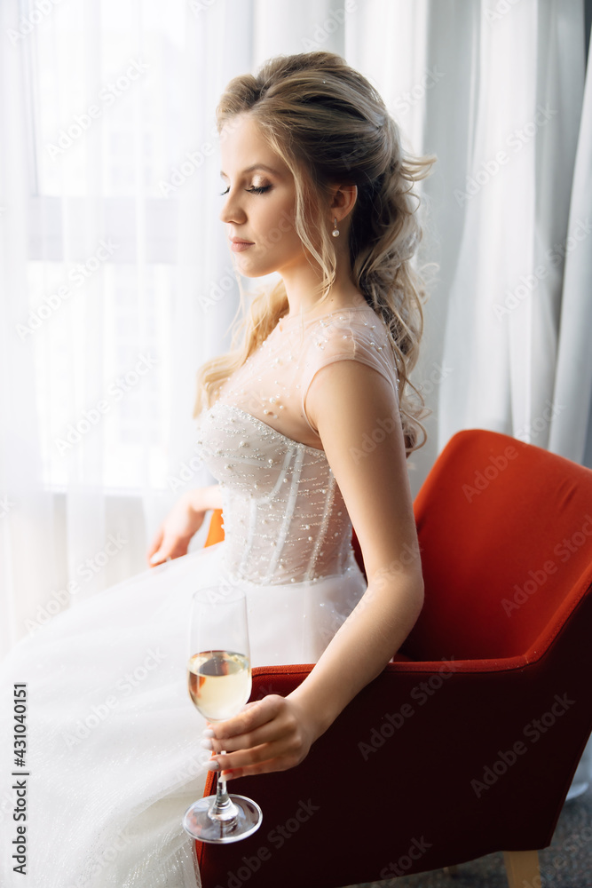 A young beautiful bride in a white wedding dress sits on a red chair with a glass of champagne. Vacation concept with drinks for adults. A young beautiful bride in a white wedding dress sits on a red chair with a glass of champagne. Vacation concept with drinks for adults.