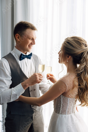 The groom in a suit and the bride in a wedding dress stand in front of the window and hold glasses of champagne drinking.Sparkling wine with bubbles and splashes on glasses.Wedding and events concept