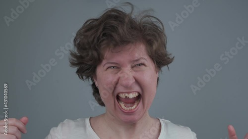 Caucasian woman is crazy grabbed pulling out her hair, lost tooth laughs and screams while looking at camera in studio on gray background. Angry edentate female tearing her hair. Mad mood photo