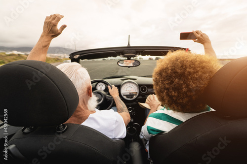Trendy senior couple having fun taking selfie with smartphone inside convertible car - Multiracial mature people on a road trip in summer vacation with cabriolet car