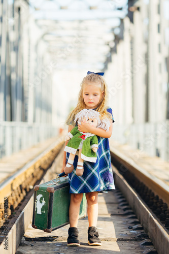 A little cute girl in a dress is waiting for her train with a suitcase at the train stations on a summer day. © Ivan