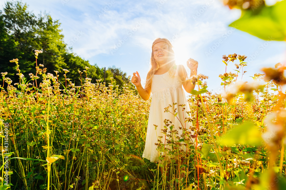 Fototapeta premium Little beautiful girl with long blond hair, walks in a yellow field, against the background of the blue sky and the sun.