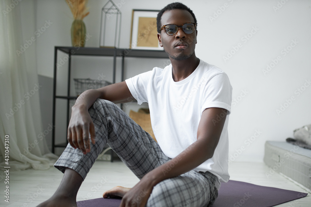 Indoor shot of serious confident young Afro American male in eyeglasses sitting barefooted on fitness mat, having rest after morning workout. People, active lifestyle, home training and domestic life