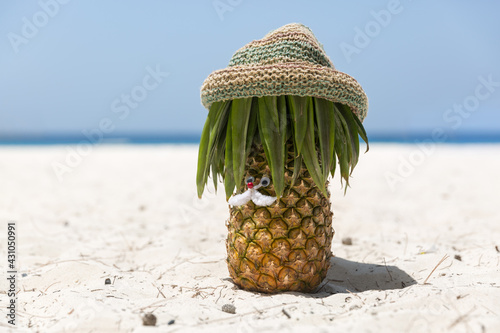 Ripe and juicy mustachioed pineapple in a hippie hat stands on the beach on white sand, in the background the azure sea and blue sky