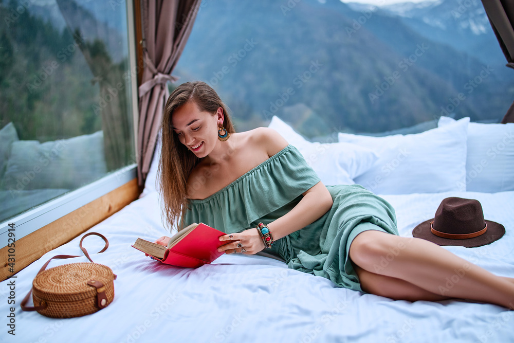 Young beautiful charming happy cute smiling woman passionate about reading exciting book while lying on bed with mountain view