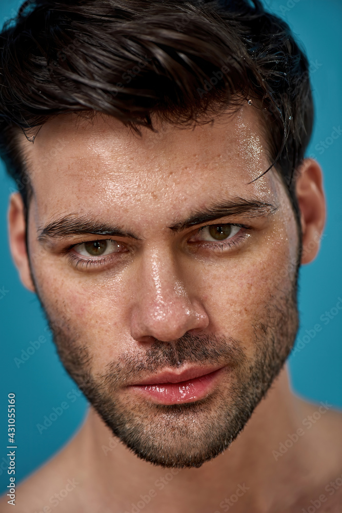 Fototapeta premium Close up portrait of good looking brunette man with wet washed face looking at camera, posing isolated over blue background