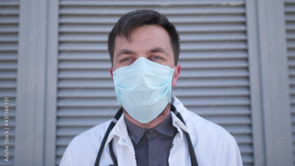Close-up of a tired male doctor removing a medical mask from his face after a heavy shift while standing outside near the wall of the hospital backyard. Concept of treatment of covid-19 corona virus Close-up of a tired male doctor removing a medical mask from his face after a heavy shift while standing outside near the wall of the hospital backyard. Concept of treatment of covid-19 corona virus