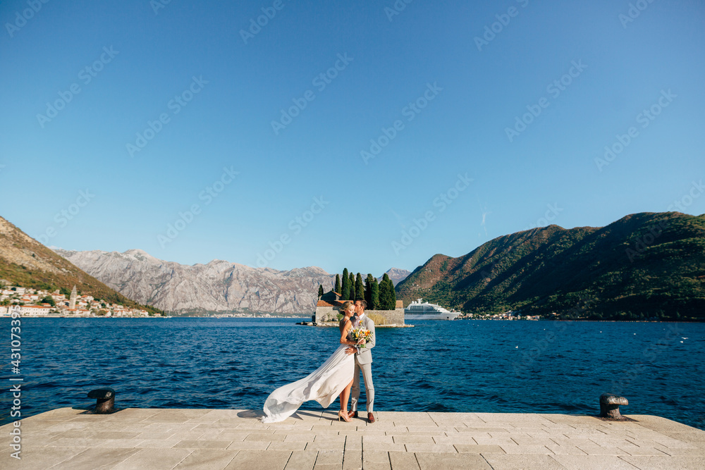 The bride and groom are embracing on the pier in the Bay of Kotor, behind them are mountains, island and old town of Perast