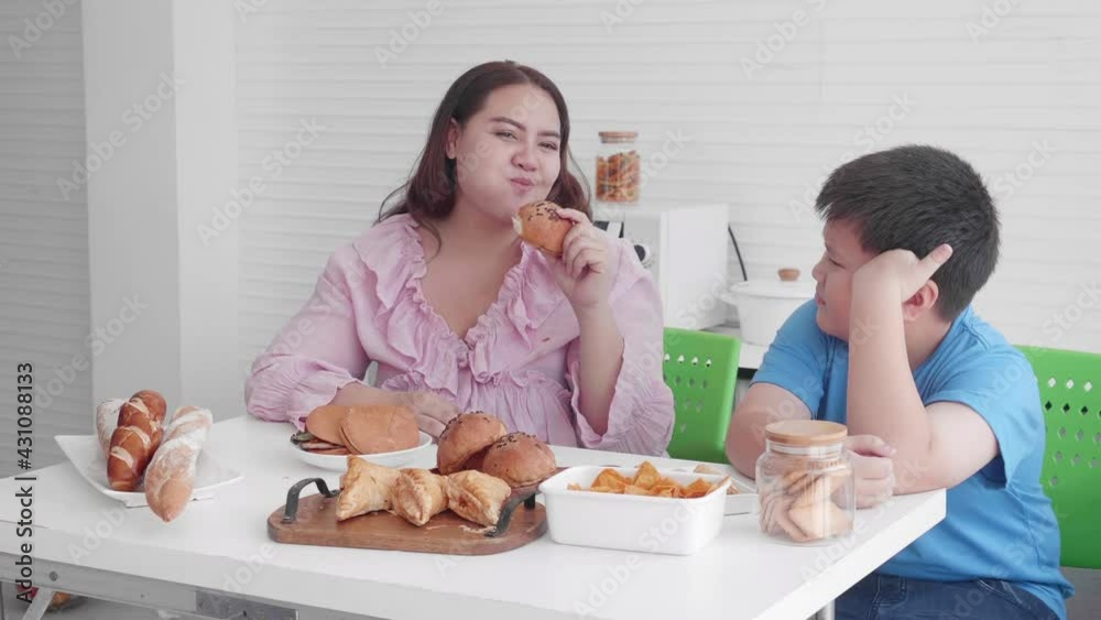 Asian family relationship of son upset with mother fat eating bread in the kitchen at home, mom hungry and eating bun with unhealthy food, two people, woman overweight, health care concept.