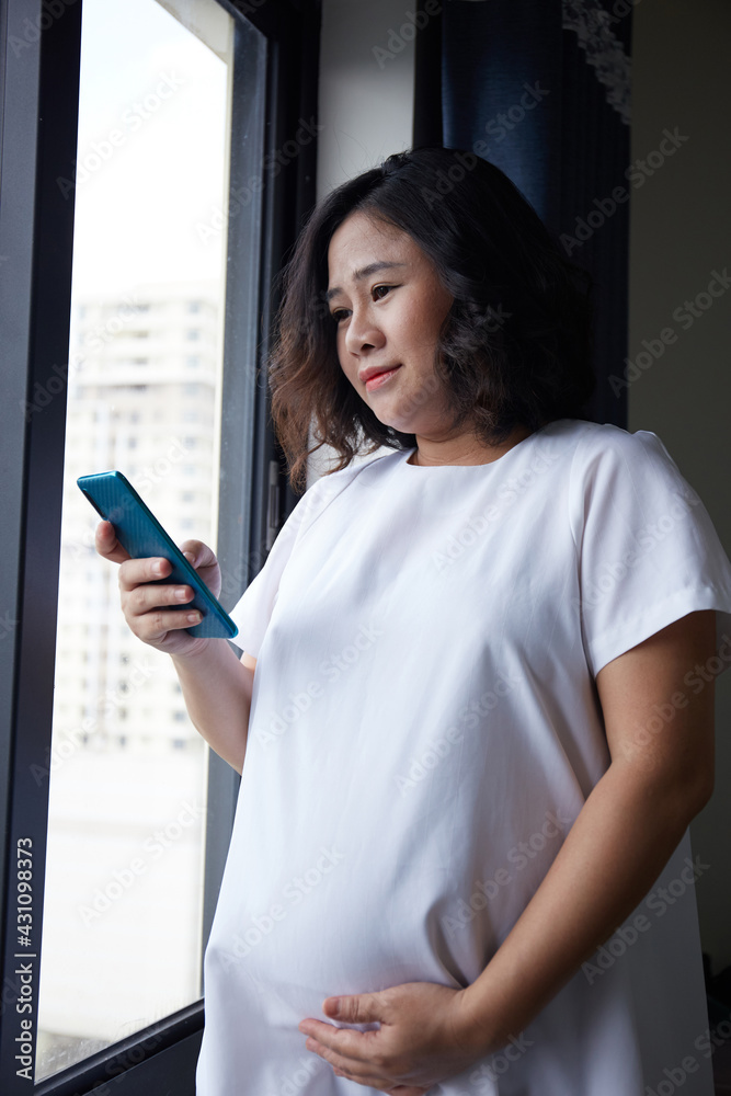 Young Asian Pregnant Woman standing near the window and using smart phone for checking information Young Asian Pregnant Woman standing near the window and using smart phone for checking information