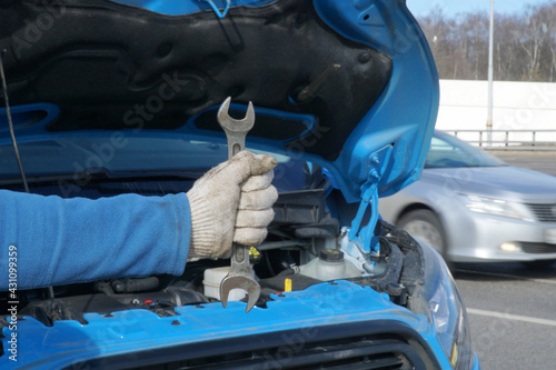 A man's hand with a wrench over a car engine with an open hood. The concept of breakdown and repair in transit.