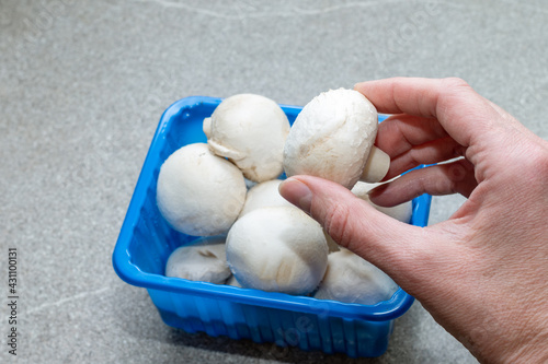 A hand takes out a Champignon Mushroom from a blue box on a gray kitchen table, close-up. Champignons white whole in a plastic box. Healthy natural food. Diet food concept