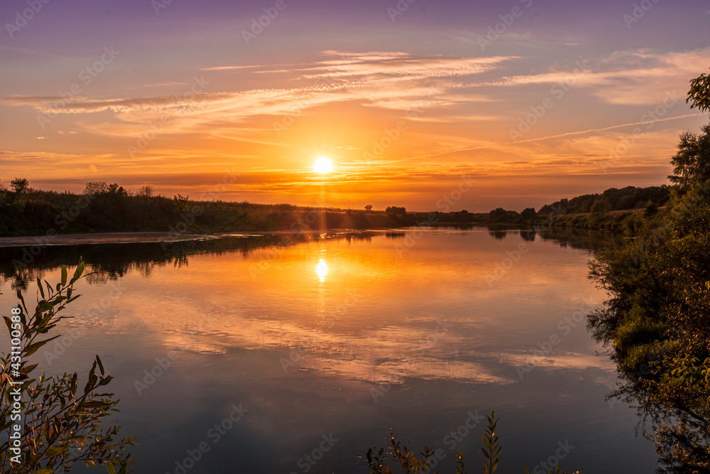 Scenic view at beautiful summer river sunset with reflection on water with green bushes, grass, golden sun rays, calm water ,deep blue cloudy sky and glow on a background, spring evening landscape