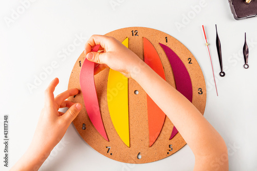 DIY and kid's creativity. Step by step instruction: how to wall clock. Step3 childs hands glue colored parts to wood pannel. Children Craft workshop. photo