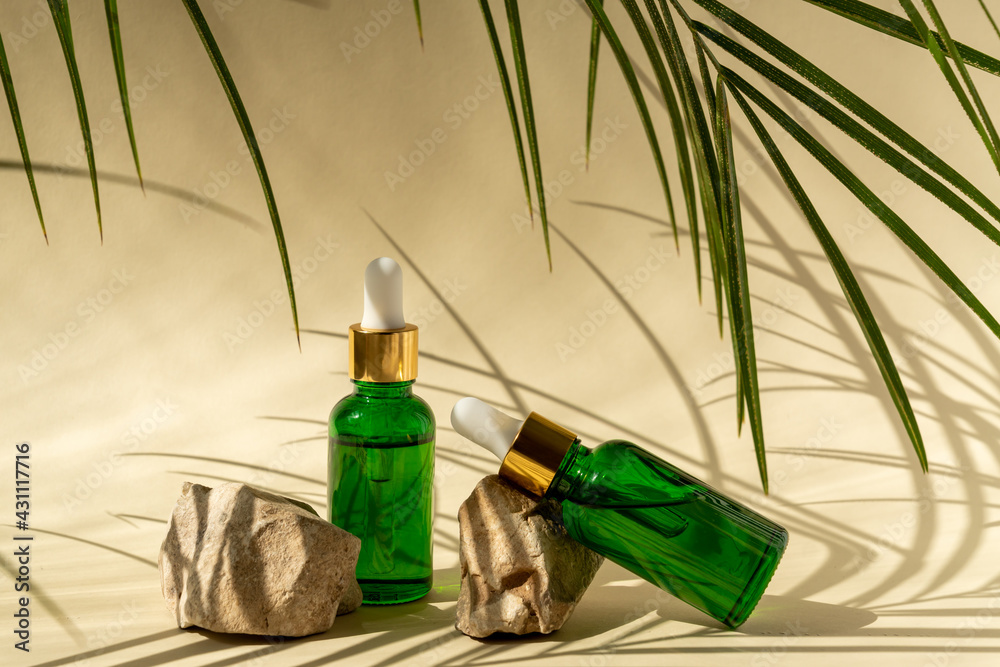 Green glass cosmetic bottles with a dropper on a beige background with stones and tropical leaves. Natural cosmetics concept, natural essential oil and skin care products Green glass cosmetic bottles with a dropper on a beige background with stones and tropical leaves. Natural cosmetics concept, natural essential oil and skin care products
