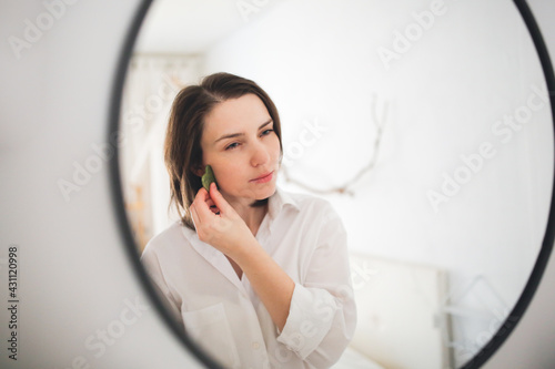 Cute caucasian woman with Jade gouache scraper near mirror ,female brunette does facial massage with scraper gouache. Concept beauty and self-care, skin and face care