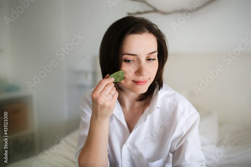 Cute caucasian woman with Jade gouache scraper in bedroom ,female brunette does facial massage with scraper gouache. Concept beauty and self-care, skin and face care