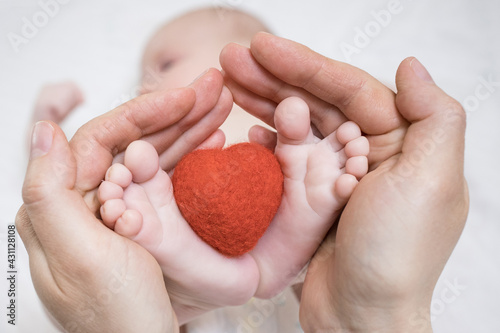 Feet of a newborn baby in mother's palms hold a small red heart. The concept of love, health and care, motherhood. 