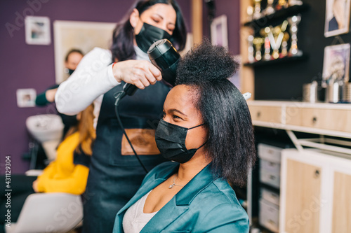 A hairdresser dries a Black woman's hair in a modern hair salon. They are both wearing face protective masks due to Coronavirus epidemic.