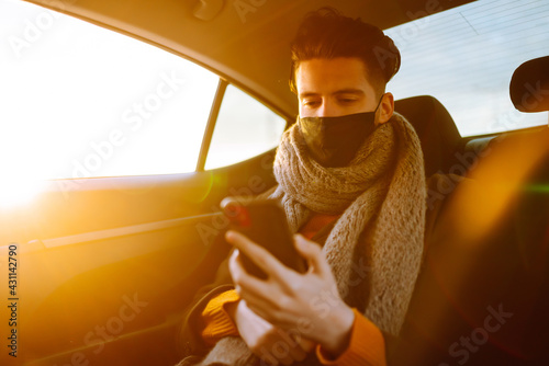 Young man passenger in protective medical mask in the taxi car on a backseat with phone. Work on the way to the office. Covid-2019.