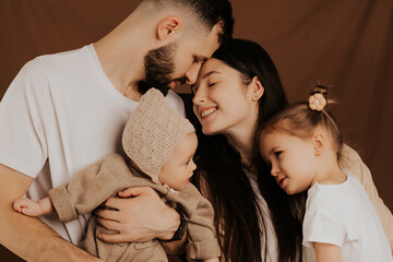 A young sincere family where a stylish bearded father and a cute wife hold their cute children in their arms. Family photo shoot in the studio