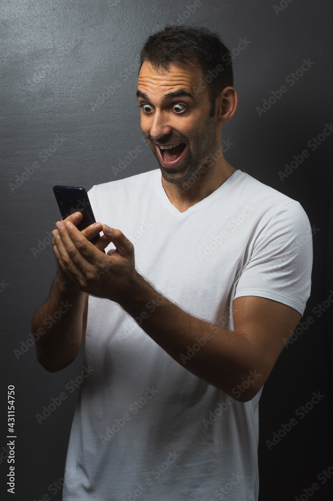 Man wearing a white tshirt over a grey background holding his phone with both hands and looking surprised Man wearing a white tshirt over a grey background holding his phone with both hands and looking surprised