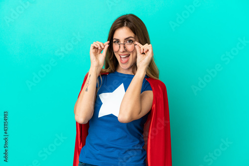 Super Hero caucasian woman isolated on blue background with glasses and surprised