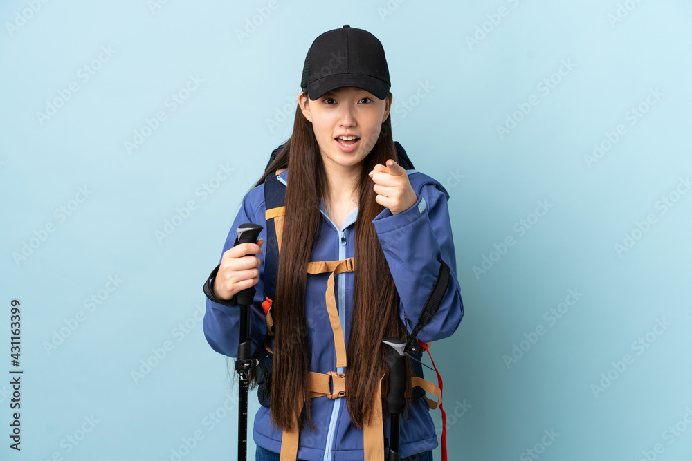 Young Chinese girl with backpack and trekking poles over isolated blue background surprised and pointing front Young Chinese girl with backpack and trekking poles over isolated blue background surprised and pointing front