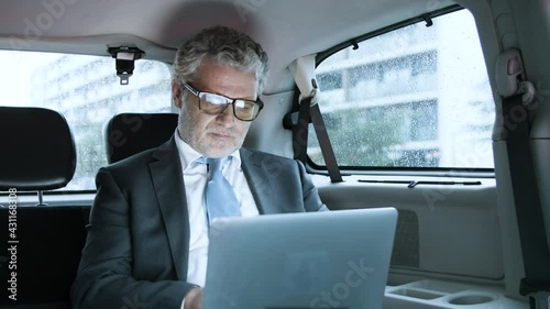 Focused senior businessman using laptop in car. Bearded man in eyeglasses and official suit typing on computer, working in car. Raining outside. Business, lifestyle, technology concept. photo