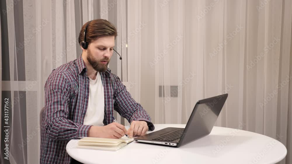 Smiling positive operator representative speaking to web camera makes note in the evening. Businessman wears headset having video conference calling at home. Customer support service call center phone Smiling positive operator representative speaking to web camera makes note in the evening. Businessman wears headset having video conference calling at home. Customer support service call center phone