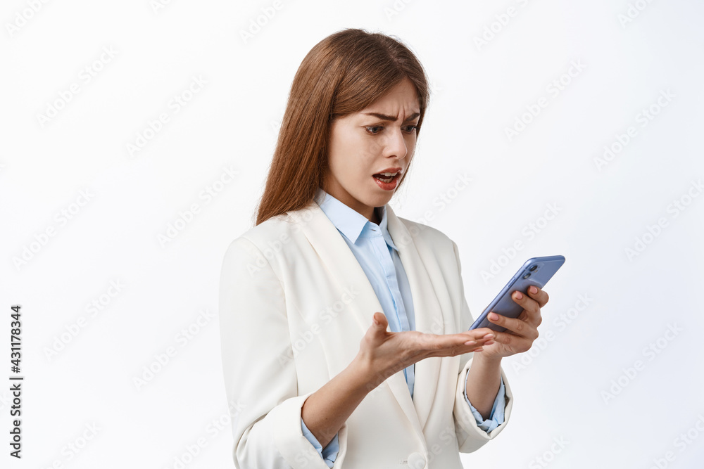 Confused woman read strange message on smartphone and complains. Business girl in corporate suit stares puzzled at phone screen, stands over white background Confused woman read strange message on smartphone and complains. Business girl in corporate suit stares puzzled at phone screen, stands over white background