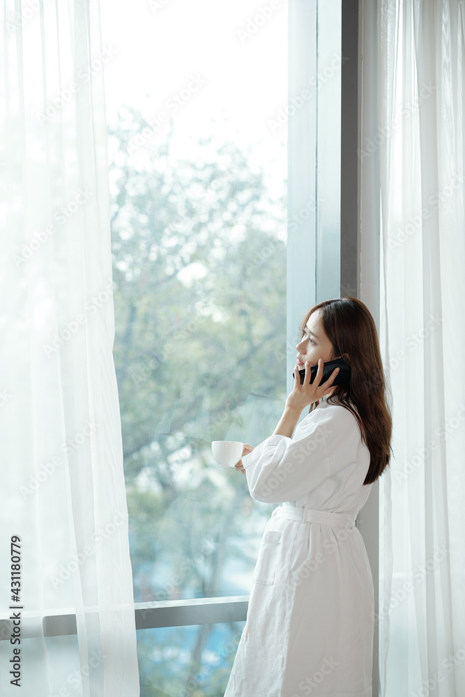 Young Asian woman in bathrobe standing at apartment window, drinking morning coffee and talking on phone Young Asian woman in bathrobe standing at apartment window, drinking morning coffee and talking on phone