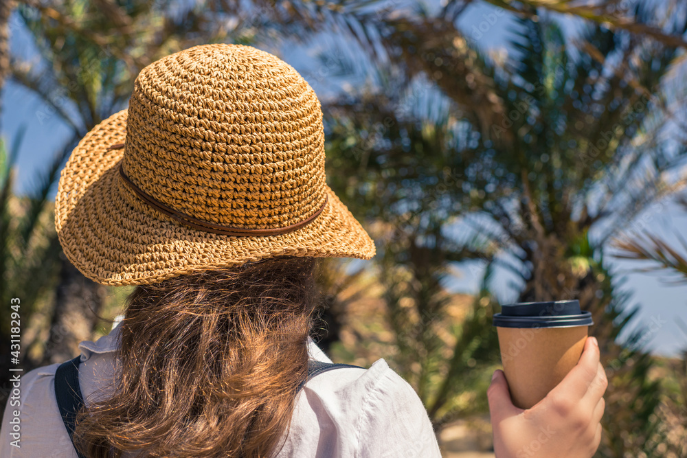 Back view on the Woman in the straw hat with paper cup of coffee walking in the palm forest.