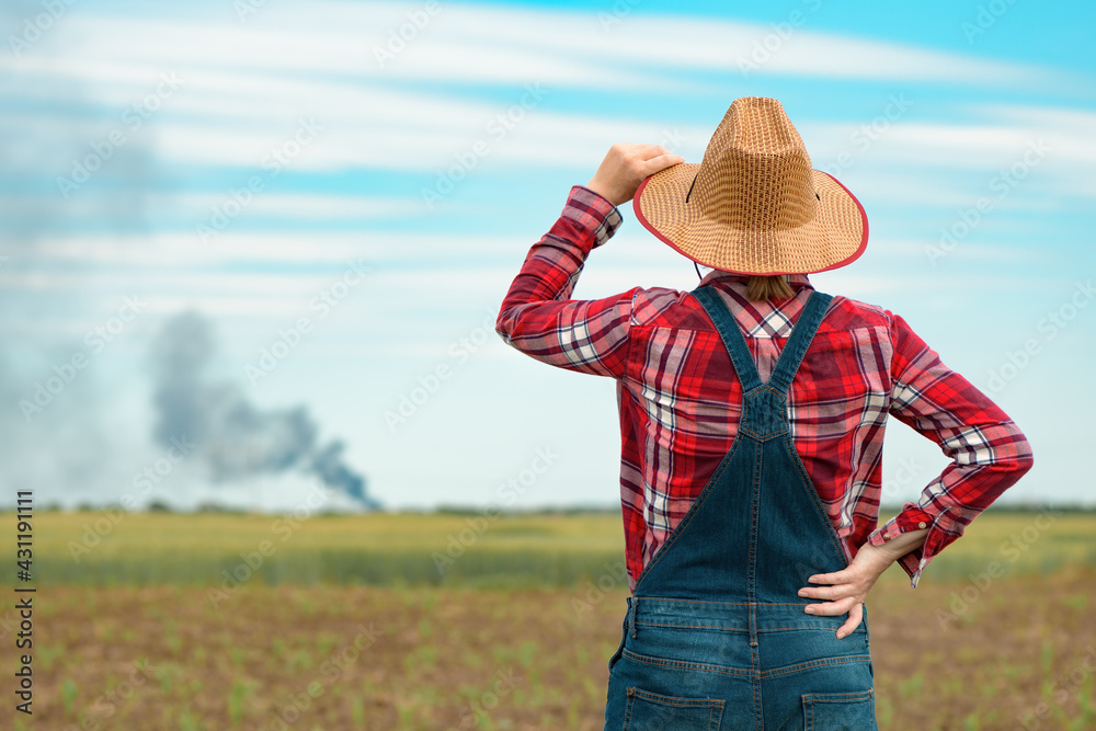 Concerned female farmer in corn field looking at black smoke on horizon