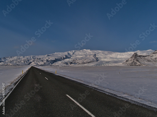 Stunning diminishing perspective of deserted, paved ring road (route 1) with markings on road trip in south Iceland in winter with the snow-covered foothills of Öræfajökull volcano and clear sky.