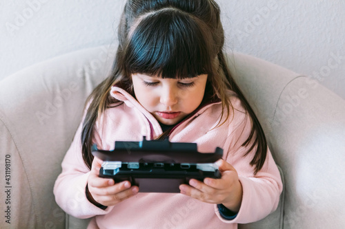 Little black haired girl wearing a pink sweater playing a video game with a portable video console on a sofa