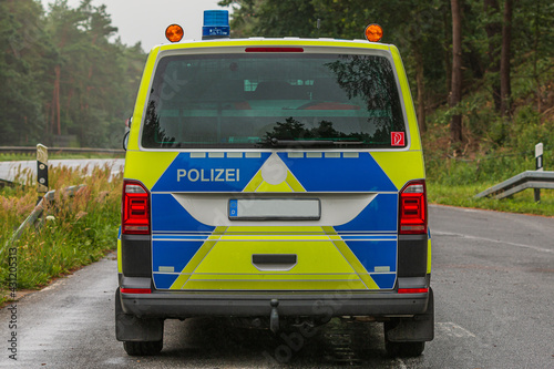 Rear view of a police car on the highway in an emergency parking bay. Rainy weather with trees in the background. Blue lights on the roof and yellow and blue paintwork
