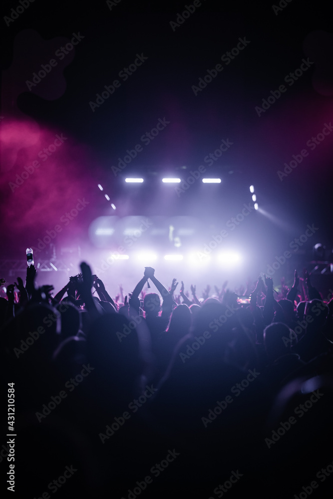Vertical shot of a cheering crowd dancing with raised hands in a party in front of a bright stage - obrazy, fototapety, plakaty 