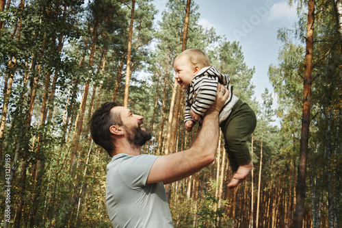 Happy father holds his baby high in the air against the backdrop of forest and tall pines.