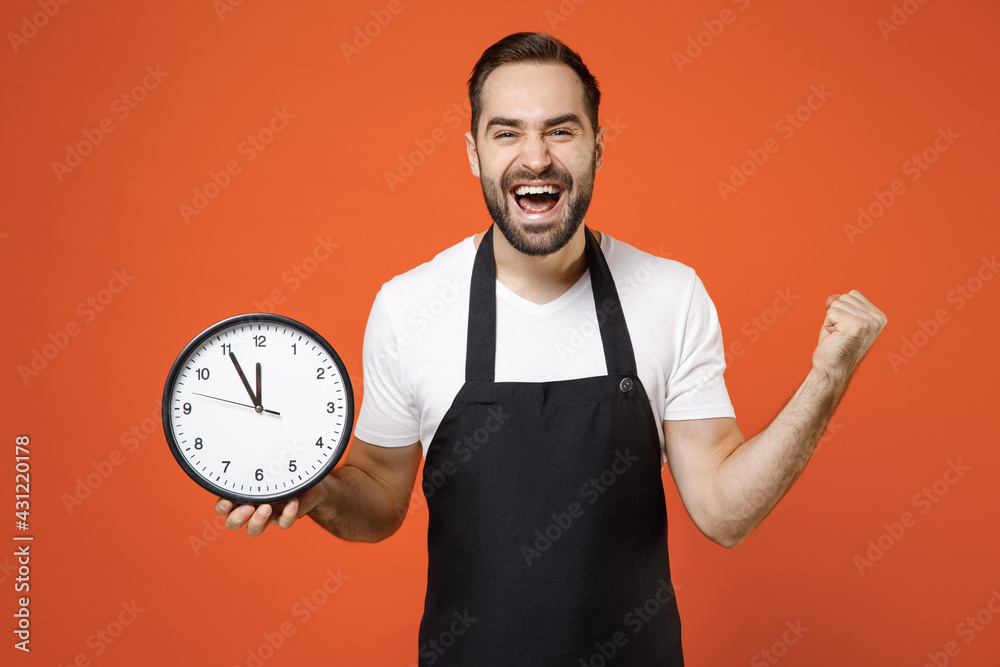 Young man barista bartender barman employee wear black apron white tshirt work in coffee shop hold clock do winner gesture isolated on orange background Small business startup time management concept Young man barista bartender barman employee wear black apron white tshirt work in coffee shop hold clock do winner gesture isolated on orange background Small business startup time management concept