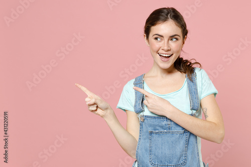 Young smiling happy cheerful caucasian woman 20s in trendy denim clothes blue t-shirt point index finger aside on workspace area mock up copy space isolated on pastel pink background studio portrait