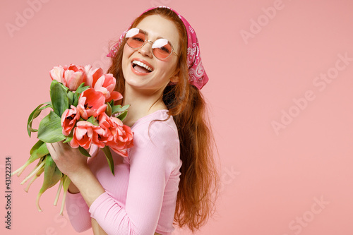 Side view of young smiling cheerful romantic woman 20s in rose clothes bandana glasses hold tulips flowers bouquet look camera isolated on pastel pink background studio portrait Spring season concept