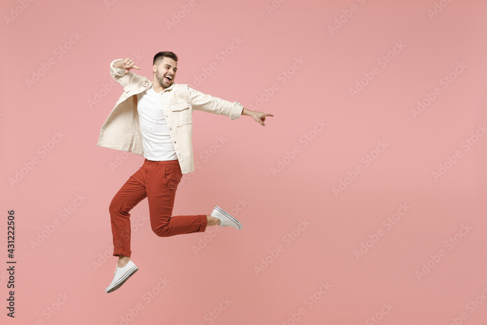 Full length young smiling excited overjoyed joyful fashionable caucasian man 20s in jacket white t-shirt jump high point index finger aside on workspace area isolated on pastel pink background studio.