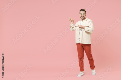 Full length young overjoyed happy trendy fashionable friendly caucasian man 20s in jacket white t-shirt point index finger aside on copy space workspace area isolated on pastel pink background studio