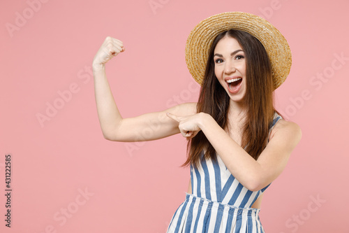 Young woman in summer clothes striped dress straw hat put arm on biceps muscles on hand demonstrating strength power isolated on pastel pink color background studio portrait. People lifestyle concept.