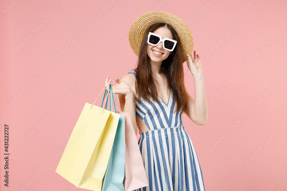 Young smiling caucasian woman 20s wear summer clothes striped dress straw glasses hold package bags with purchases after shopping touch hat look camera isolated on pastel pink color background studio.