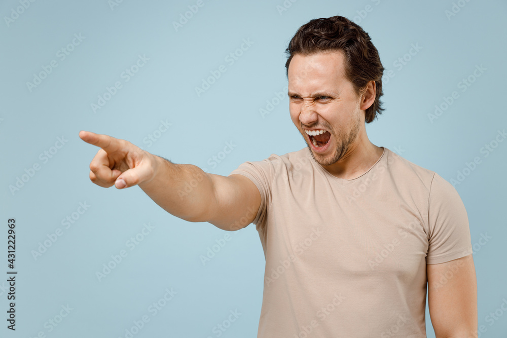 Young sad angry strict caucasian man wear casual basic beige t-shirt point index finger aside scream shout do it isolated on pastel blue color background studio portrait. People lifestyle concept Young sad angry strict caucasian man wear casual basic beige t-shirt point index finger aside scream shout do it isolated on pastel blue color background studio portrait. People lifestyle concept